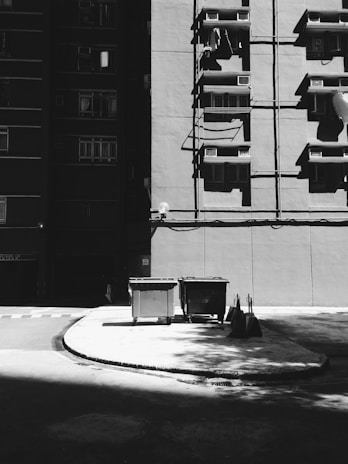 A black and white image of a building facade with several windows and air conditioning units. Shadows cast across the concrete ground, and two large garbage bins are situated near the building. A few broomsticks and dustpans are leaning against the curb.