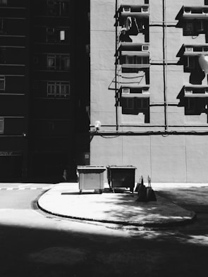 A black and white image of a building facade with several windows and air conditioning units. Shadows cast across the concrete ground, and two large garbage bins are situated near the building. A few broomsticks and dustpans are leaning against the curb.