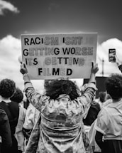 A group of people are gathered with one person holding a sign overhead that reads 'RACISM ISNT GETTING WORSE ITS GETTING FILMED'. The scene captures a moment of protest or demonstration. The image is in black and white, adding a stark and serious tone to the context.