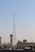 A large metal communication tower stands prominently against a clear blue sky, with a cityscape visible in the background. The tower is tall and is surrounded by various buildings, including one with a distinctive spherical structure on top.