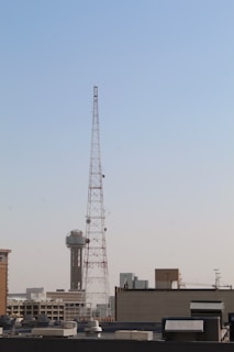 A large metal communication tower stands prominently against a clear blue sky, with a cityscape visible in the background. The tower is tall and is surrounded by various buildings, including one with a distinctive spherical structure on top.
