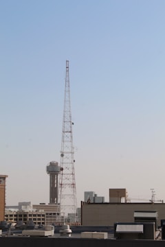 A large metal communication tower stands prominently against a clear blue sky, with a cityscape visible in the background. The tower is tall and is surrounded by various buildings, including one with a distinctive spherical structure on top.