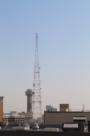 A large metal communication tower stands prominently against a clear blue sky, with a cityscape visible in the background. The tower is tall and is surrounded by various buildings, including one with a distinctive spherical structure on top.
