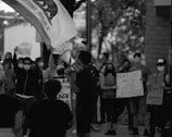 A group of people is gathered in protest, holding signs with messages like 'Black Lives Matter' and 'I Can’t Breathe.' Many individuals are wearing masks, and one person is speaking into a megaphone. A California Republic flag is visible, adding context to the location.