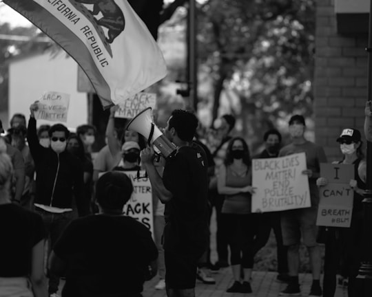 A group of people is gathered in protest, holding signs with messages like 'Black Lives Matter' and 'I Can’t Breathe.' Many individuals are wearing masks, and one person is speaking into a megaphone. A California Republic flag is visible, adding context to the location.