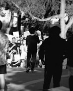 A black and white photo depicts a crowd gathered outdoors, likely for a protest or demonstration. A person holding a megaphone stands with their back to the camera. Some participants are seated, while others stand, many holding signs. Facial expressions range from serious to attentive, with a mixture of masked and unmasked individuals.