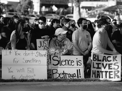 A black and white photograph of a group of people gathered at a protest. Several individuals are holding signs with messages advocating for justice and awareness. The participants are wearing face masks, suggesting pandemic precautions. The scene conveys a sense of solidarity and activism.