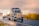 A large blue semi-truck with a long flatbed trailer travels on a wet road. The trailer is carrying white, bagged cargo. The surroundings are vibrant with autumn foliage and the sky is overcast, giving a moody yet serene atmosphere with hints of sunshine breaking through.