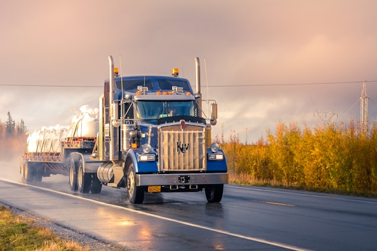 A large blue semi-truck with a long flatbed trailer travels on a wet road. The trailer is carrying white, bagged cargo. The surroundings are vibrant with autumn foliage and the sky is overcast, giving a moody yet serene atmosphere with hints of sunshine breaking through.