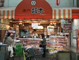 A small shop with a striped orange and white awning, displaying a variety of meat products in glass cases. Two people, likely customers, stand at the counter where a shopkeeper is present. Bicycles and a shopping cart are in the foreground.