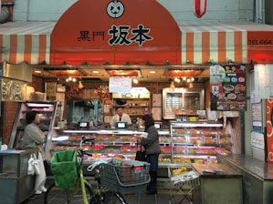 A small shop with a striped orange and white awning, displaying a variety of meat products in glass cases. Two people, likely customers, stand at the counter where a shopkeeper is present. Bicycles and a shopping cart are in the foreground.