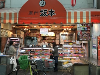 A small shop with a striped orange and white awning, displaying a variety of meat products in glass cases. Two people, likely customers, stand at the counter where a shopkeeper is present. Bicycles and a shopping cart are in the foreground.