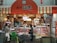 A small shop with a striped orange and white awning, displaying a variety of meat products in glass cases. Two people, likely customers, stand at the counter where a shopkeeper is present. Bicycles and a shopping cart are in the foreground.