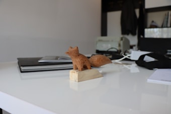 A small wooden fox sculpture sits on a smooth white surface. In the background, there are books and electronic devices, giving a home office or studio-like atmosphere. The fox is intricately carved and stands on a rectangular base.