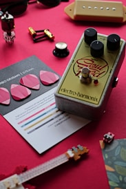 A collection of musical accessories and electronics are arranged on a red surface. The focal point is a guitar effects pedal with knobs labeled volume, drive, and treble, and the brand name Electro-Harmonix. Surrounding the pedal are various objects including guitar picks, a small brochure, a hardware component resembling a vacuum tube, and several small components resembling switches and tuning pegs. The arrangement appears to be a curated flat lay related to music and instruments.