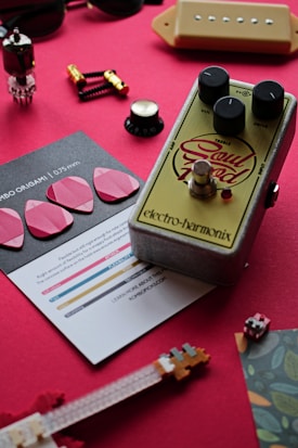A collection of musical accessories and electronics are arranged on a red surface. The focal point is a guitar effects pedal with knobs labeled volume, drive, and treble, and the brand name Electro-Harmonix. Surrounding the pedal are various objects including guitar picks, a small brochure, a hardware component resembling a vacuum tube, and several small components resembling switches and tuning pegs. The arrangement appears to be a curated flat lay related to music and instruments.