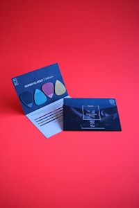 Two promotional cards against a bright red background. One card displays colorful guitar picks and has a modern design with text detailing the thickness and features. The other features a more subdued design with an image of a guitar and text about the product's new generation.