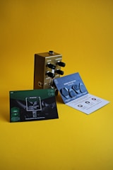 A metallic guitar effects pedal with black knobs labeled for volume, compression, release, and attack is placed on a yellow surface. In front of the pedal, there are two promotional cards—one green and one blue with text and images of guitar picks.