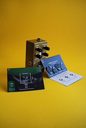 A metallic guitar effects pedal with black knobs labeled for volume, compression, release, and attack is placed on a yellow surface. In front of the pedal, there are two promotional cards—one green and one blue with text and images of guitar picks.
