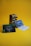 A metallic guitar effects pedal with black knobs labeled for volume, compression, release, and attack is placed on a yellow surface. In front of the pedal, there are two promotional cards—one green and one blue with text and images of guitar picks.