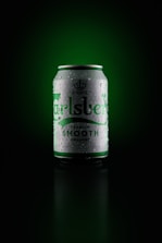 A can of Carlsberg beer is centered against a dark green background. The can is silver with green lettering and is covered in droplets, giving it a cold, refreshing appearance.