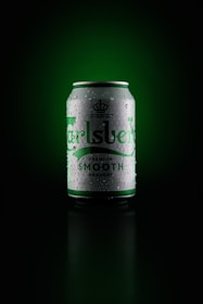 A can of Carlsberg beer is centered against a dark green background. The can is silver with green lettering and is covered in droplets, giving it a cold, refreshing appearance.