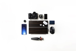 A flat lay of various electronic gadgets and accessories on a white background includes a camera, smartphone, wireless earbuds, computer mouse, portable hard drive, camera lens, and a tripod. A tablet with a dark cover is positioned centrally, accompanied by a lens cap.