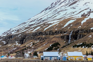 A mountainous landscape featuring snow-capped peaks and rolling hills. A small town with colorful houses, including blue and yellow, is situated at the base of the mountains. A waterfall runs down the rocky surface, bordered by patches of greenery.