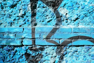 A textured blue wall featuring a mix of cobblestones and bricks. The surface is painted with an intricate graffiti design in black, creating a striking contrast with the vivid blue background.