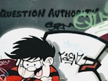 A graffiti art piece on a wall depicts a cartoon character with spiky black hair and a mischievous expression, wearing a red and black striped shirt. The words 'Question Authority' are boldly painted above, surrounded by various tags and abstract designs in green and black.
