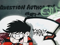 A graffiti art piece on a wall depicts a cartoon character with spiky black hair and a mischievous expression, wearing a red and black striped shirt. The words 'Question Authority' are boldly painted above, surrounded by various tags and abstract designs in green and black.