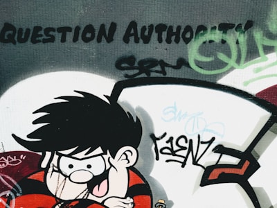 A graffiti art piece on a wall depicts a cartoon character with spiky black hair and a mischievous expression, wearing a red and black striped shirt. The words 'Question Authority' are boldly painted above, surrounded by various tags and abstract designs in green and black.