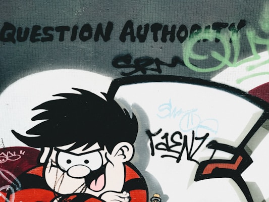 A graffiti art piece on a wall depicts a cartoon character with spiky black hair and a mischievous expression, wearing a red and black striped shirt. The words 'Question Authority' are boldly painted above, surrounded by various tags and abstract designs in green and black.