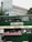 A green double-decker bus has been repurposed as a food truck, offering tacos, burritos, and tortas. The setup includes a colorful tablecloth with various condiments and menus displayed. The bus is parked in an urban area, with modern buildings and greenery visible in the background.