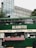 A green double-decker bus has been repurposed as a food truck, offering tacos, burritos, and tortas. The setup includes a colorful tablecloth with various condiments and menus displayed. The bus is parked in an urban area, with modern buildings and greenery visible in the background.