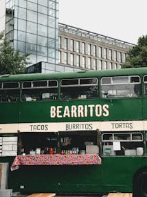 A green double-decker bus has been repurposed as a food truck, offering tacos, burritos, and tortas. The setup includes a colorful tablecloth with various condiments and menus displayed. The bus is parked in an urban area, with modern buildings and greenery visible in the background.
