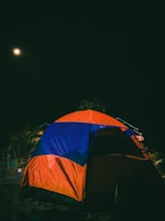 A brightly colored tent in orange and blue is set up against a dark night sky. The moon shines in the upper left corner, casting a soft glow. Nearby are some trees and vegetation, partially illuminated by the moonlight.