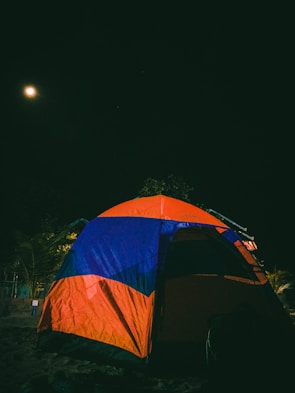 A brightly colored tent in orange and blue is set up against a dark night sky. The moon shines in the upper left corner, casting a soft glow. Nearby are some trees and vegetation, partially illuminated by the moonlight.