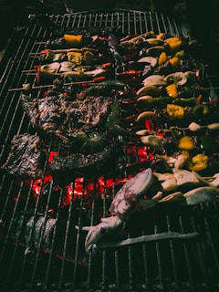 A variety of food items are being grilled on a barbecue. Vegetables and seafood, including squid, are skewered and sizzling over hot coals. Some peppers and other vegetables are charred, while the flame adds a warm glow.