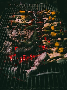A variety of food items are being grilled on a barbecue. Vegetables and seafood, including squid, are skewered and sizzling over hot coals. Some peppers and other vegetables are charred, while the flame adds a warm glow.