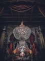 A large, ornate chandelier dominates the upper portion of the image, hanging in an intricately decorated room. Below, a statue of Ganesha is adorned with garlands, positioned at the center of an elaborately designed altar. Rich, dark drapery frames the scene, complemented by heavy architectural details.