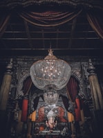 A large, ornate chandelier dominates the upper portion of the image, hanging in an intricately decorated room. Below, a statue of Ganesha is adorned with garlands, positioned at the center of an elaborately designed altar. Rich, dark drapery frames the scene, complemented by heavy architectural details.
