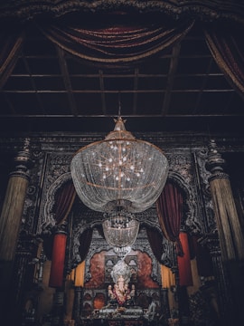 A large, ornate chandelier dominates the upper portion of the image, hanging in an intricately decorated room. Below, a statue of Ganesha is adorned with garlands, positioned at the center of an elaborately designed altar. Rich, dark drapery frames the scene, complemented by heavy architectural details.