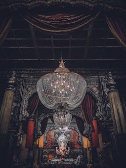 A large, ornate chandelier dominates the upper portion of the image, hanging in an intricately decorated room. Below, a statue of Ganesha is adorned with garlands, positioned at the center of an elaborately designed altar. Rich, dark drapery frames the scene, complemented by heavy architectural details.