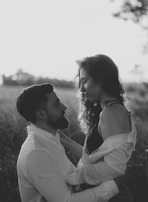 A couple is gazing into each other's eyes in a field during what appears to be a sunset. The man, with a beard, is wearing a light-colored shirt and is positioned slightly lower, suggestively kneeling or sitting. The woman, with long curly hair, is standing or sitting on his lap, wearing a black top with an open light shirt over it. The background is out of focus, emphasizing the couple, and the lighting creates a romantic silhouette effect.