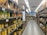 A supermarket aisle is filled with shelves stocked with various products in cardboard packaging. The shelves are labeled 'No Brand' and prices are displayed in a foreign currency. The products include cans and boxes, with some signage in a non-English language. Fluorescent lights illuminate the aisle, and a person can be seen in the distance.