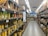 A supermarket aisle is filled with shelves stocked with various products in cardboard packaging. The shelves are labeled 'No Brand' and prices are displayed in a foreign currency. The products include cans and boxes, with some signage in a non-English language. Fluorescent lights illuminate the aisle, and a person can be seen in the distance.