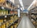 A supermarket aisle is filled with shelves stocked with various products in cardboard packaging. The shelves are labeled 'No Brand' and prices are displayed in a foreign currency. The products include cans and boxes, with some signage in a non-English language. Fluorescent lights illuminate the aisle, and a person can be seen in the distance.