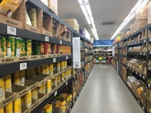 A supermarket aisle is filled with shelves stocked with various products in cardboard packaging. The shelves are labeled 'No Brand' and prices are displayed in a foreign currency. The products include cans and boxes, with some signage in a non-English language. Fluorescent lights illuminate the aisle, and a person can be seen in the distance.