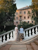 An older couple stands on a white stone balustrade, gazing at a three-story residential building. The scene is set amidst lush greenery, with tall trees partially obscuring the building. The warm lighting suggests a late afternoon or early evening setting.